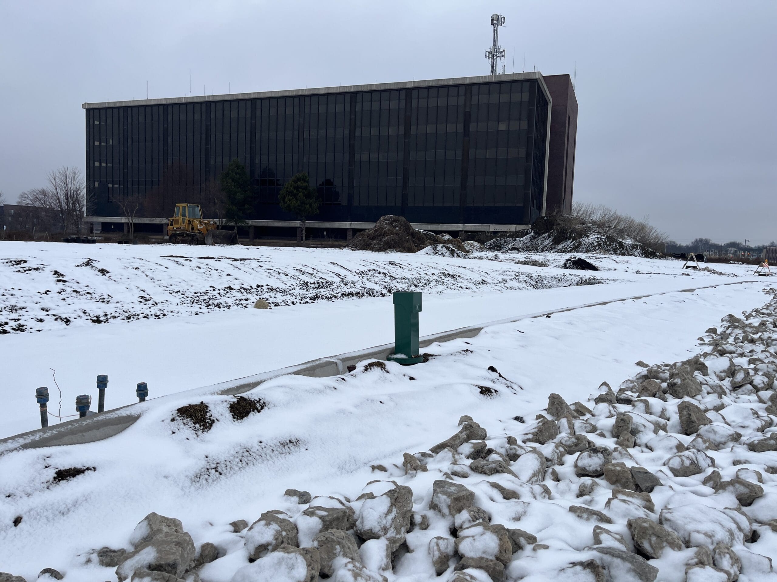A snow-covered construction site featuring a green Pedoc power pedestal foundation mounting bracket, utility connections, and a partially completed pathway. A large modern office building and construction equipment are visible in the background.